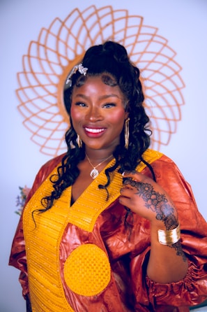 A woman stands confidently, adorned in a vibrant traditional dress featuring a combination of yellow and brown fabric. Her hair is styled in long curls, accessorized with a decorative clip. She is smiling warmly, wearing large hoop earrings and a necklace. A detailed henna design is visible on her hand, complemented by gold bangles on her wrist. Behind her, a decorative circular pattern adds to the composition.