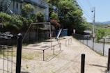 An outdoor playground area with a swing set and a slide surrounded by greenery. A fence borders the playground on one side, and there's a road visible in the background. Nearby, a few buildings are partially seen, with trees and bushes adding natural elements to the scene.
