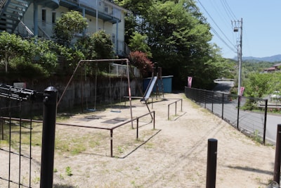 An outdoor playground area with a swing set and a slide surrounded by greenery. A fence borders the playground on one side, and there's a road visible in the background. Nearby, a few buildings are partially seen, with trees and bushes adding natural elements to the scene.