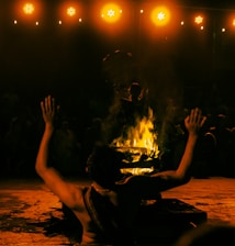 A person performs a ritual in front of a burning fire, with arms raised in the air. The scene is set in dim lighting with an audience in the background, creating a dramatic and intense atmosphere. Warm, glowing lights hang overhead, casting an orange hue on the scene.