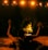 A person performs a ritual in front of a burning fire, with arms raised in the air. The scene is set in dim lighting with an audience in the background, creating a dramatic and intense atmosphere. Warm, glowing lights hang overhead, casting an orange hue on the scene.