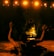 A person performs a ritual in front of a burning fire, with arms raised in the air. The scene is set in dim lighting with an audience in the background, creating a dramatic and intense atmosphere. Warm, glowing lights hang overhead, casting an orange hue on the scene.