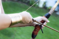 A person is drawing a bow with an arrow. The hands are adorned with metallic bracelets. The background is primarily green, suggesting an outdoor setting.