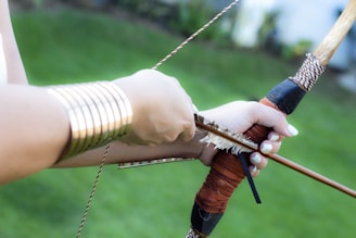 A person is drawing a bow with an arrow. The hands are adorned with metallic bracelets. The background is primarily green, suggesting an outdoor setting.