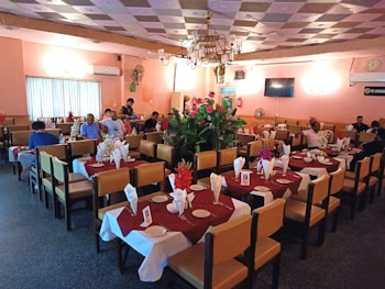 A cozy restaurant dining area with neatly set tables covered in maroon and white tablecloths. The tables are arranged in rows and adorned with folded napkins and red flowers. Several people are seated and engaged in conversation, while others appear to be waiting for their meal. The room features soft lighting, a decorated ceiling, and a chandelier in the center. Walls painted in pastel colors add to the relaxed atmosphere.