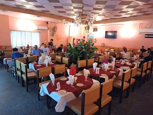 A cozy restaurant dining area with neatly set tables covered in maroon and white tablecloths. The tables are arranged in rows and adorned with folded napkins and red flowers. Several people are seated and engaged in conversation, while others appear to be waiting for their meal. The room features soft lighting, a decorated ceiling, and a chandelier in the center. Walls painted in pastel colors add to the relaxed atmosphere.