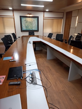 A modern conference room with a large, oval wooden table surrounded by black office chairs. The table has a white strip in the center, with a few electronic devices and cables on it, including a laptop and remote controls. The room is decorated with a large abstract painting on the wall and is illuminated by overhead lights. Wooden floors and blinds on the windows add to the contemporary look.