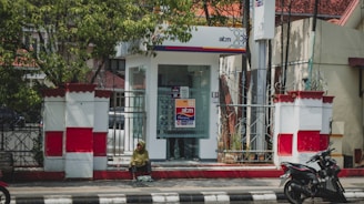 A small ATM booth with glass walls is situated on a walkway, surrounded by decorative white and red fencing. A person sits on the curb in the foreground, dressed in a green top with a headscarf, while a motorcycle is parked to the right.