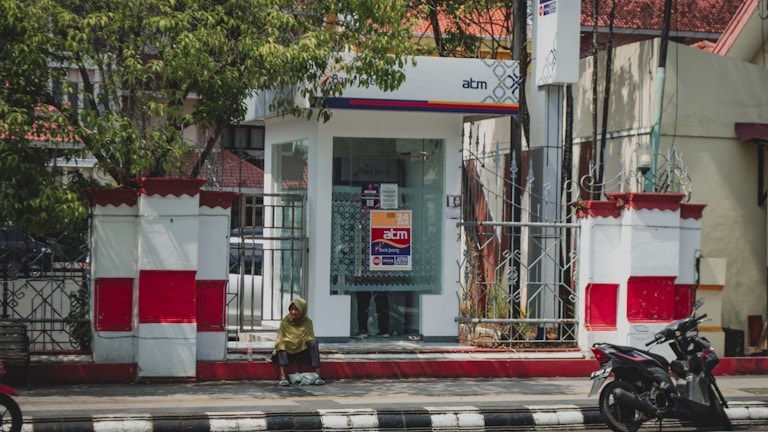 A small ATM booth with glass walls is situated on a walkway, surrounded by decorative white and red fencing. A person sits on the curb in the foreground, dressed in a green top with a headscarf, while a motorcycle is parked to the right.