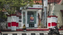 A small ATM booth with glass walls is situated on a walkway, surrounded by decorative white and red fencing. A person sits on the curb in the foreground, dressed in a green top with a headscarf, while a motorcycle is parked to the right.
