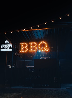A dimly lit outdoor setting with a prominent sign that reads 'BBQ' illuminated in warm, amber lights. The area seems to be a food stall or small restaurant specializing in barbeque, as indicated by the word 'BARBEQUE' on a smaller illuminated sign nearby. The ambiance is cozy and inviting, with string lights adding a touch of charm above.