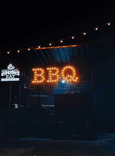 A dimly lit outdoor setting with a prominent sign that reads 'BBQ' illuminated in warm, amber lights. The area seems to be a food stall or small restaurant specializing in barbeque, as indicated by the word 'BARBEQUE' on a smaller illuminated sign nearby. The ambiance is cozy and inviting, with string lights adding a touch of charm above.