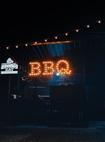 A dimly lit outdoor setting with a prominent sign that reads 'BBQ' illuminated in warm, amber lights. The area seems to be a food stall or small restaurant specializing in barbeque, as indicated by the word 'BARBEQUE' on a smaller illuminated sign nearby. The ambiance is cozy and inviting, with string lights adding a touch of charm above.