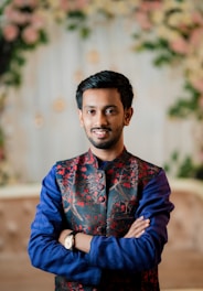 A person stands with arms crossed, wearing a traditional embroidered jacket with red floral patterns over a blue kurta. The background is softly blurred with green and pink floral elements.