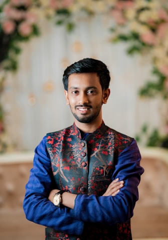 A person stands with arms crossed, wearing a traditional embroidered jacket with red floral patterns over a blue kurta. The background is softly blurred with green and pink floral elements.