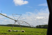 A large irrigation system spans a vast field of green crops under a bright, partly cloudy sky. The equipment is positioned to water the plants evenly, with its metal structure extending across the landscape. Trees are visible in the distance, marking the edge of the field.