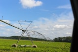 A large irrigation system spans a vast field of green crops under a bright, partly cloudy sky. The equipment is positioned to water the plants evenly, with its metal structure extending across the landscape. Trees are visible in the distance, marking the edge of the field.
