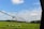 A large irrigation system spans a vast field of green crops under a bright, partly cloudy sky. The equipment is positioned to water the plants evenly, with its metal structure extending across the landscape. Trees are visible in the distance, marking the edge of the field.