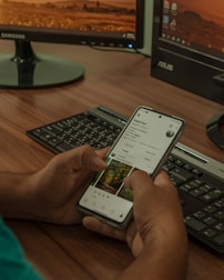 A person holding a smartphone displaying a social media profile, with two computer monitors and a keyboard in the background. The setting appears to be a workspace with a wooden desk.