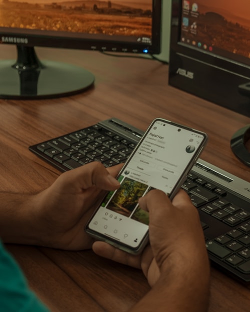 A person holding a smartphone displaying a social media profile, with two computer monitors and a keyboard in the background. The setting appears to be a workspace with a wooden desk.