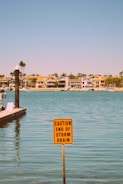 A calm body of water with a sign in the foreground warning about the end of a storm drain. The background features several modern houses lining the shoreline under a clear blue sky.