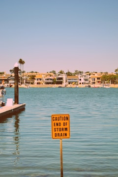 A calm body of water with a sign in the foreground warning about the end of a storm drain. The background features several modern houses lining the shoreline under a clear blue sky.