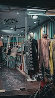 A clothing store with a mannequin in a traditional black outfit displayed at the entrance. Inside, several people are interacting, possibly customers and staff. The walls are adorned with multi-colored vertical stripes. There are various traditional garments hanging inside. The atmosphere seems busy and lively, with bright lighting in the store.