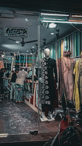 A clothing store with a mannequin in a traditional black outfit displayed at the entrance. Inside, several people are interacting, possibly customers and staff. The walls are adorned with multi-colored vertical stripes. There are various traditional garments hanging inside. The atmosphere seems busy and lively, with bright lighting in the store.