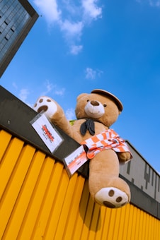 A large teddy bear wearing a hat and an orange and white checkered piece of clothing is perched atop a yellow corrugated wall under a clear blue sky. Part of a tall building is visible in the corner, and there are some signs attached to the wall next to the teddy bear.