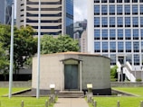 A small, round concrete building with a metal door is situated on a neatly maintained lawn. Tall, modern skyscrapers can be seen in the background, along with lush green trees. The sky is partly cloudy, and there is a sense of urban tranquility.