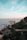 High-rise residential buildings are situated near the coastline, with the sea visible in the background. A lush green hillside borders the buildings, showing a mix of dense trees and rocky terrain. The sky above is expansive with soft blue and hints of pastel hues.