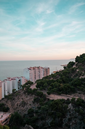 High-rise residential buildings are situated near the coastline, with the sea visible in the background. A lush green hillside borders the buildings, showing a mix of dense trees and rocky terrain. The sky above is expansive with soft blue and hints of pastel hues.