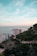High-rise residential buildings are situated near the coastline, with the sea visible in the background. A lush green hillside borders the buildings, showing a mix of dense trees and rocky terrain. The sky above is expansive with soft blue and hints of pastel hues.