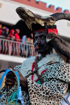 A person is adorned in a detailed, traditional outfit, featuring a headdress with large horns and animal skin patterns. An eagle is perched on the individual's shoulder, symbolizing a connection with nature or tradition. The background shows a gathering of people, suggesting a festival or cultural event.