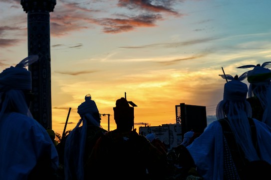The image captures a group of people silhouetted against a vibrant sunset, dressed in traditional garments with headdresses. The background features a tall, ornate column, and there's an overall festive atmosphere.