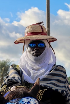 A person is wearing traditional clothing that includes a large woven hat with red accents and a face covering. They have reflective sunglasses, and the background features a partly cloudy sky. The attire suggests cultural or regional significance, possibly indicating a festival or traditional event.
