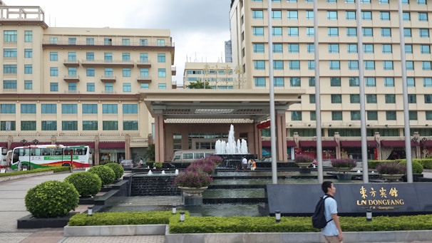 A multi-story hotel building with a fountain in front, surrounded by manicured gardens and pathways. Buses are parked on the side, and a person walks past the entrance where a sign displays the name of the establishment.