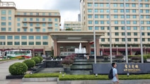 A multi-story hotel building with a fountain in front, surrounded by manicured gardens and pathways. Buses are parked on the side, and a person walks past the entrance where a sign displays the name of the establishment.