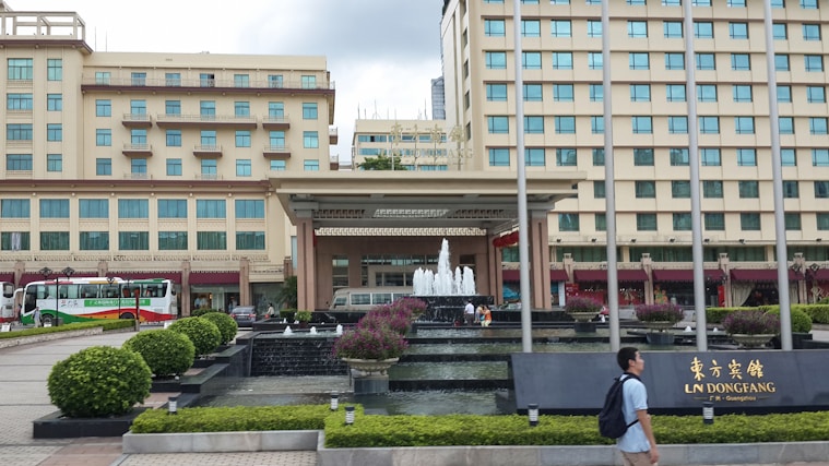 A multi-story hotel building with a fountain in front, surrounded by manicured gardens and pathways. Buses are parked on the side, and a person walks past the entrance where a sign displays the name of the establishment.