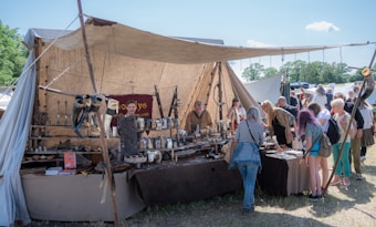 A market stall displays various medieval-style goods, including drinking horns, pottery, and metal crafts. The stall is covered by a beige canvas and has a rustic appearance. Several people are gathered around, examining the merchandise. The background shows more tents and trees under a bright blue sky.