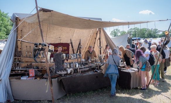 A market stall displays various medieval-style goods, including drinking horns, pottery, and metal crafts. The stall is covered by a beige canvas and has a rustic appearance. Several people are gathered around, examining the merchandise. The background shows more tents and trees under a bright blue sky.