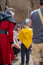 A group of people is gathered in a historical setting. A man in a red and black costume stands on the left, while a child in a yellow cloak and helmet is in the middle. Another man in casual attire is on the right, and two women are sitting or crouching in the background, engaged in a conversation.