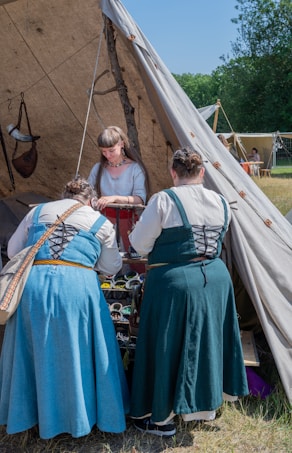 Three women in period clothing are gathered around a tent. Two women in blue dresses are facing the tent while another woman in a light dress is standing inside the tent, working on something. The tent is set outdoors in a grassy area with more tents visible in the background. The weather appears to be clear and sunny.