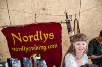 A young woman with earrings and a necklace is smiling, seated in front of a red cloth banner with the text 'Nordlys nordlysviking.com.' Behind her, there is a rustic setting with wooden and metallic objects resembling Viking-themed items.