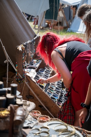 A person with bright red hair is looking through a display of colorful beads and handmade jewelry at an outdoor market stall. The stall has various items such as necklaces, bracelets, and bowls of beads. The setup is rustic, with wooden planks and canvas tents visible in the background.