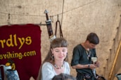 Two people dressed in historical or medieval-style clothing are inside a tent. A woman is in the foreground, wearing earrings and a colorful necklace, with her hair styled uniquely. In the background, a man is focused on something he's holding, possibly a drinking horn. Behind them is a burgundy tapestry with text.
