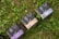Several boxes with elegant designs labeled with different product names are placed on a patch of green grass and clover. The boxes have distinct typography and color schemes, including shades of purple, blue, and beige.