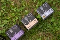 Several boxes with elegant designs labeled with different product names are placed on a patch of green grass and clover. The boxes have distinct typography and color schemes, including shades of purple, blue, and beige.