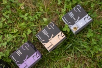 Several boxes with elegant designs labeled with different product names are placed on a patch of green grass and clover. The boxes have distinct typography and color schemes, including shades of purple, blue, and beige.