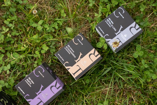Several boxes with elegant designs labeled with different product names are placed on a patch of green grass and clover. The boxes have distinct typography and color schemes, including shades of purple, blue, and beige.